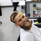 Mann sitzt auf dem Schreibtischstuhl und lacht in die Kamera. Auf seiner Stirn klebt ein Zettel mit der Aufschrift: Be Happy.