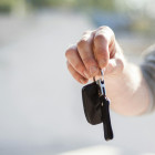 Eine Person hält dem Betrachter einen Autoschlüssel am ausgestreckten Arm entgegen.