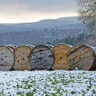 Fünf Glasfaserkabeltrommeln liegen in einer Reihe vor waldigen Hügeln auf einem Feld im Schnee.