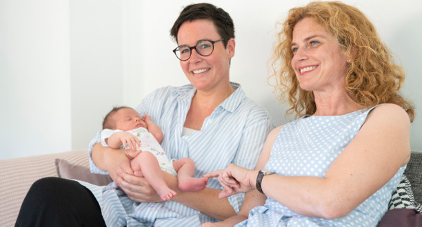 Zwei Frauen sitzen auf einem Sofa. Eine von ihnen hält ein Baby in ihrem Arm.