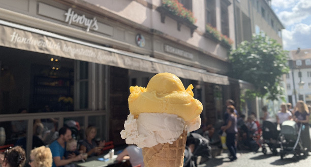 Ein Eis wird hochgehalten. Im Hintergrund ist eine Eisdiele zu sehen.