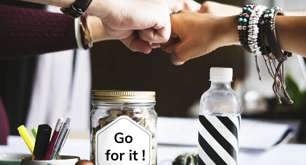 Drei Fäuste, die zusammen über einem Tisch kommen. Im Vordergrund steht ein Glas mit der Aufschrift: Go for it!