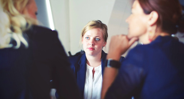 Drei Frauen sitzen an einem Tisch. Eine von diesen schaut nach vorne. Die anderen beiden sitzen mit dem Rücken zur Kamera.