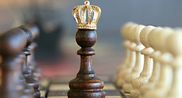 Schachfigur mit einer Krone. Links stehen braune und rechts weiße Figuren.