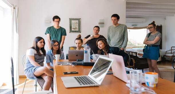 Viele Menschen stehen und sitzen um einen Tisch mit Laptops.