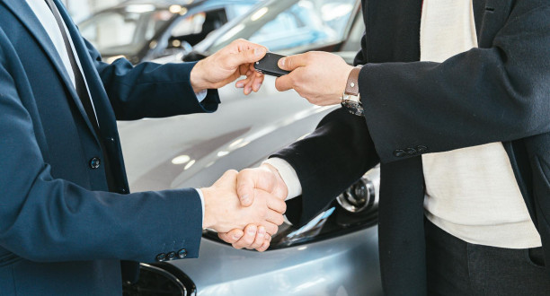 Eine Person überreicht mit der linken Hand einer anderen Person einen Autoschlüssel. Beide schütteln sich die rechte Hand.