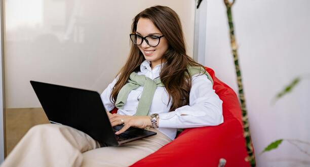 Frau mit Brille sitzt auf einem roten Stuhl mit Laptop auf dem Schoß.