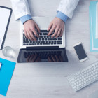 Ein Arzt sitzt an einem Tisch mit Block, Laptop und Stethoskop