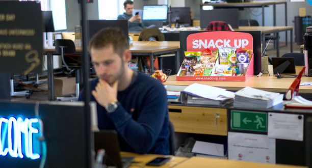 Ein Mann sitzt vor seinem PC im Hintergrund ist eine Box mit Snacks zu sehen.
