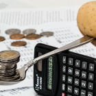 Ein Löffel liegt auf einem Taschenrechner. Auf diesem liegen vorne Münzen und am Ende eine Kartoffel.