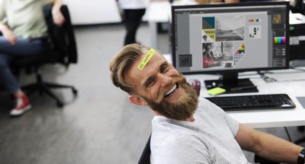 Mann sitzt auf dem Schreibtischstuhl und lacht in die Kamera. Auf seiner Stirn klebt ein Zettel mit der Aufschrift: Be Happy.