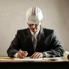 Ein Mann mit einem weißen Helm sitzt am Tisch. Vor ihm Dokumente, die er unterzeichnet.
