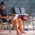 Mann sitzt mit Urlaubsoutfit auf Sonnenstuhl und hält Laptop auf Schoß.