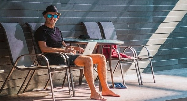 Mann sitzt mit Urlaubsoutfit auf Sonnenstuhl und hält Laptop auf Schoß.