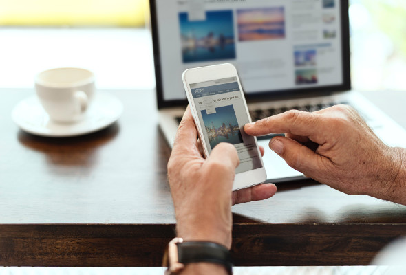 Mann hält ein Handy in der Hand.Im Hintergrund steht ein Laptop