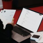 Eine Person sitzt mit einem Laptop und Papieren an einem Schreibtisch.