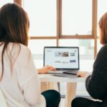 Zwei Frauen sitzen an einem Laptop, auf dem Display ist ein Onlineshop sichtbar.