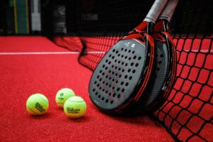 Drei Padel-Bälle liegen vor zwei Padel-Schlägern, welche an einem Padel-Netz auf einem Padel-Platz lehnen.