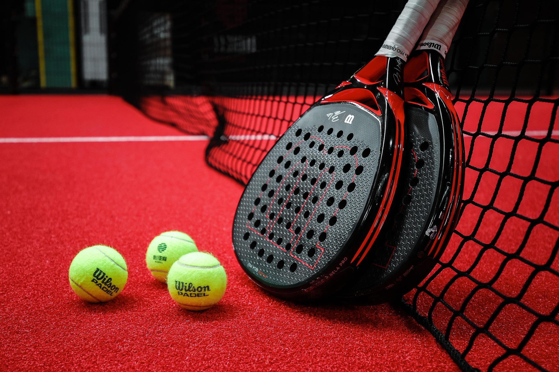 Drei Padel-Bälle liegen vor zwei Padel-Schlägern, welche an einem Padel-Netz auf einem Padel-Platz lehnen.